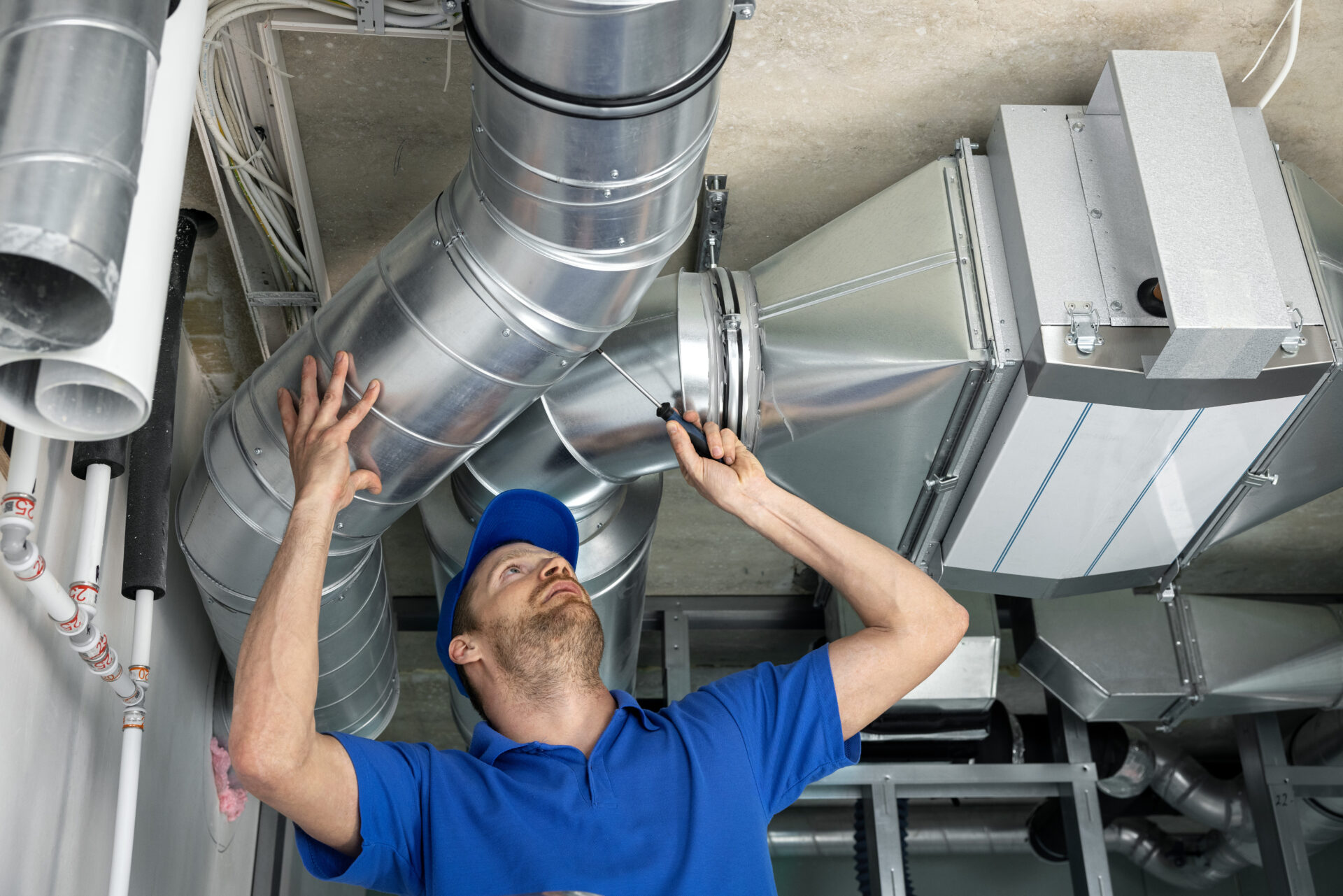 A man in a blue short-sleeved shirt and hat inspects shiny air ducts