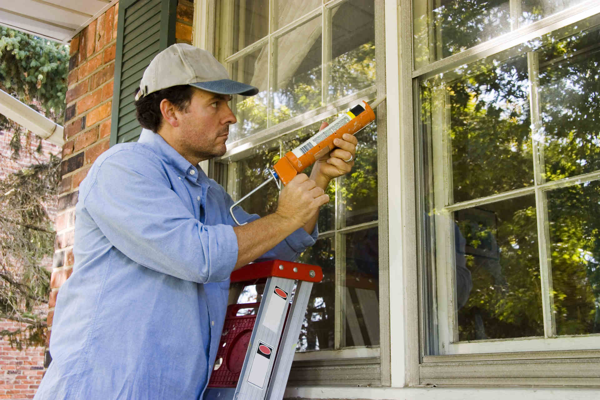 dark-haired man in a blue button-down shirt and ball cap on a ladder caulking the exterior of a window