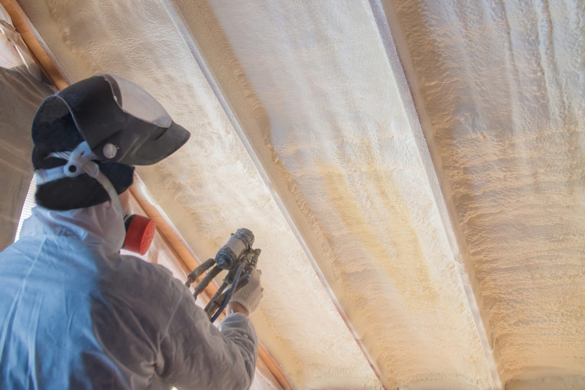 person in white protective gear and a black helmet installing spray foam insulation on a ceiling