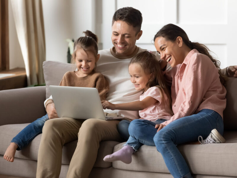 happy dark-haired family with two young kids smiling on a couch looking at a laptop screen