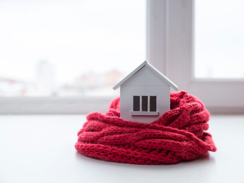 model of a white house with three windows wrapped in a red knit scarf on a white tabletop with a window in the background