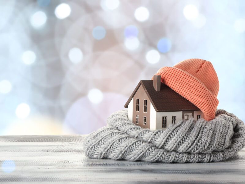 model of a house wrapped in a gray scarf and terracotta colored hat on a wood surface with a blurred background