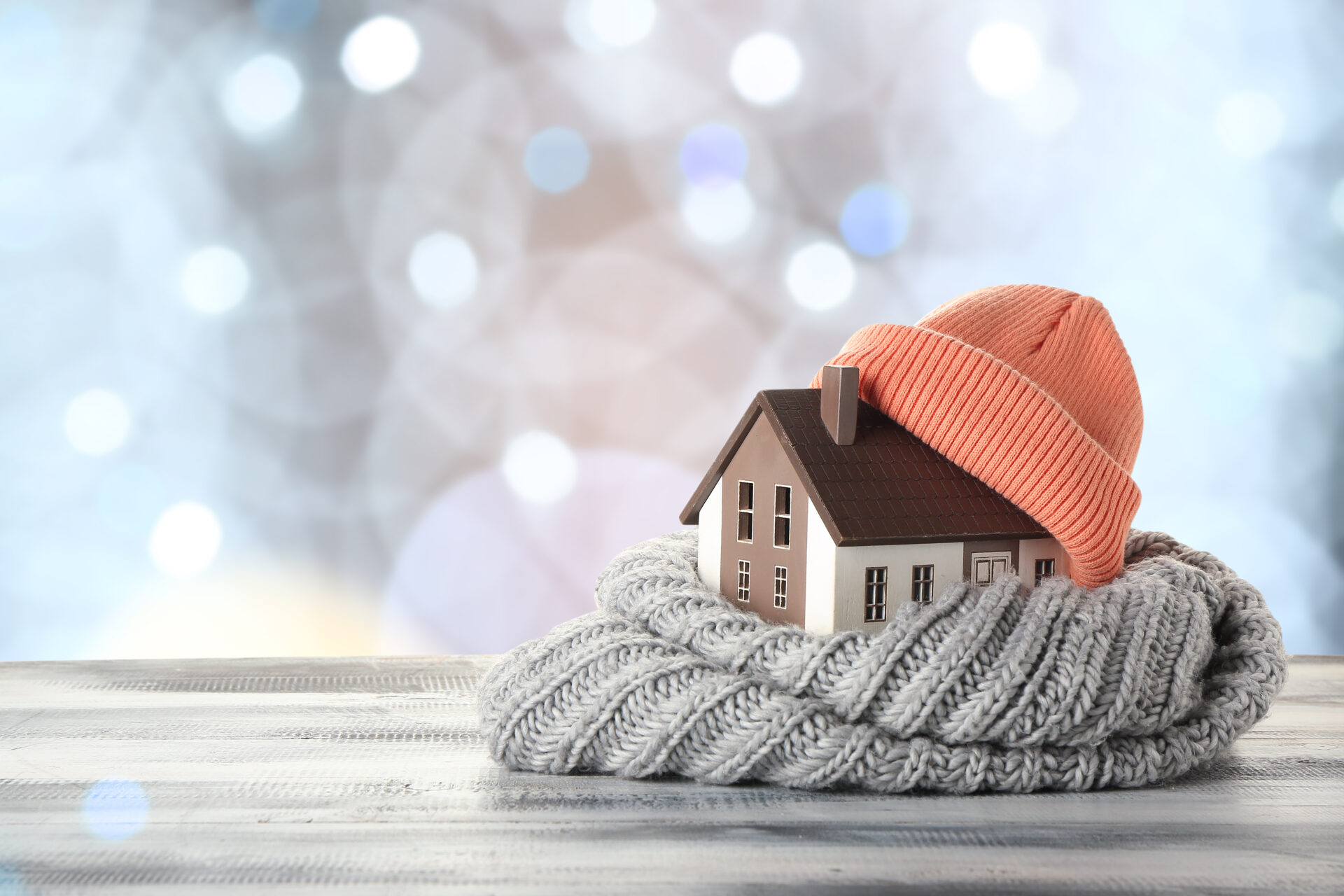 model of a house wrapped in a gray scarf and terracotta colored hat on a wood surface with a blurred background