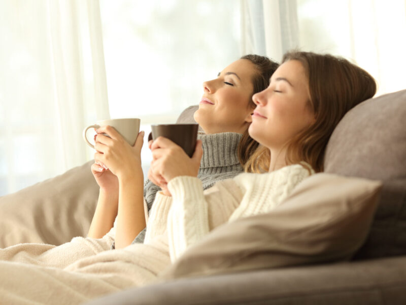 two dark-haired women sit side by side holding coffee cups and smiling contentedly indicating comfort and relaxation