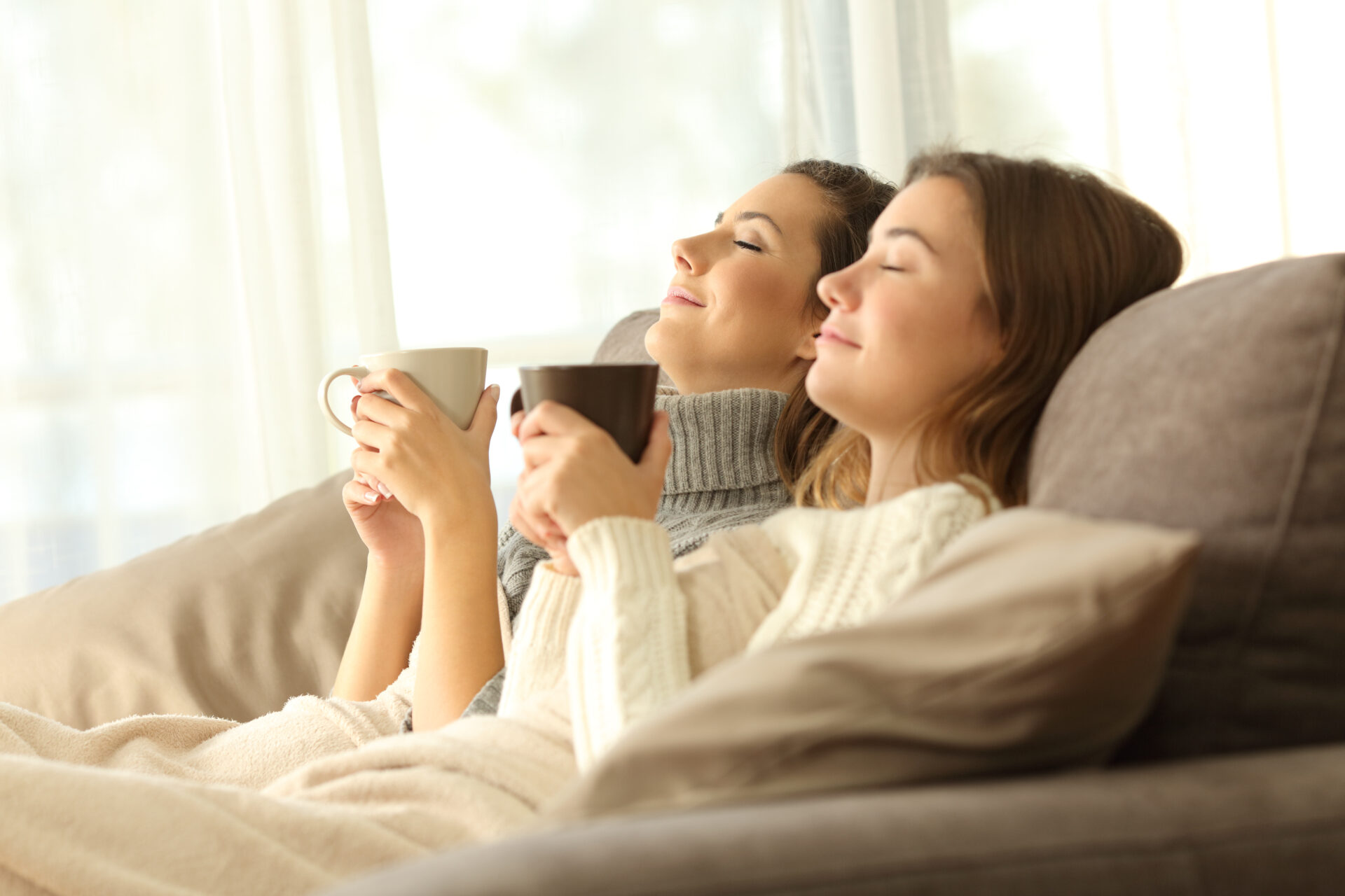 two dark-haired women sit side by side holding coffee cups and smiling contentedly indicating comfort and relaxation