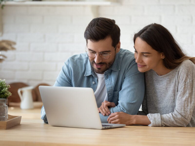 a smiling couple surf the internet, researching available NYS programs on a silver laptop set on a wooden countertop in a room with white brick walls