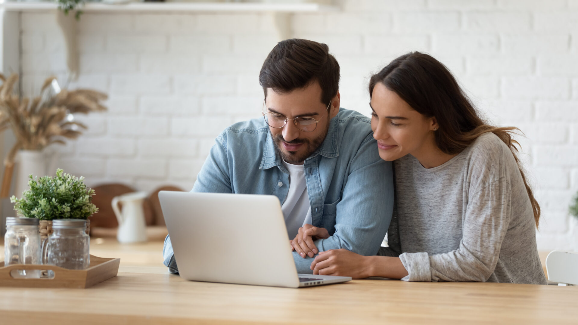 a smiling couple surf the internet, researching available NYS programs on a silver laptop set on a wooden countertop in a room with white brick walls