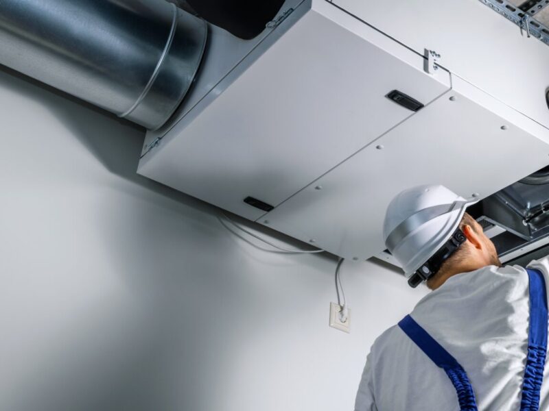 A homeowner wearing a white hardhat, white T-shirt, and blue overalls checks his HVAC system