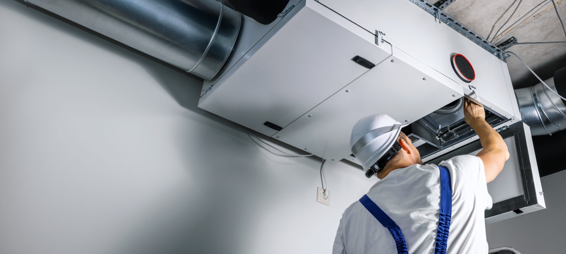 A homeowner wearing a white hardhat, white T-shirt, and blue overalls checks his HVAC system