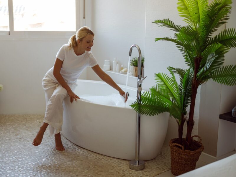 A blond woman wearing a white shirt and pants draws a bath, happy with the temperature of the water