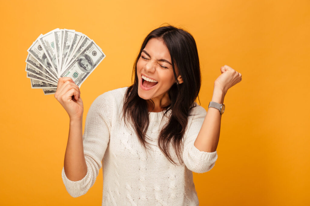 A smiling dark-haired woman in a white shirt holds a fanned-out pile of money 