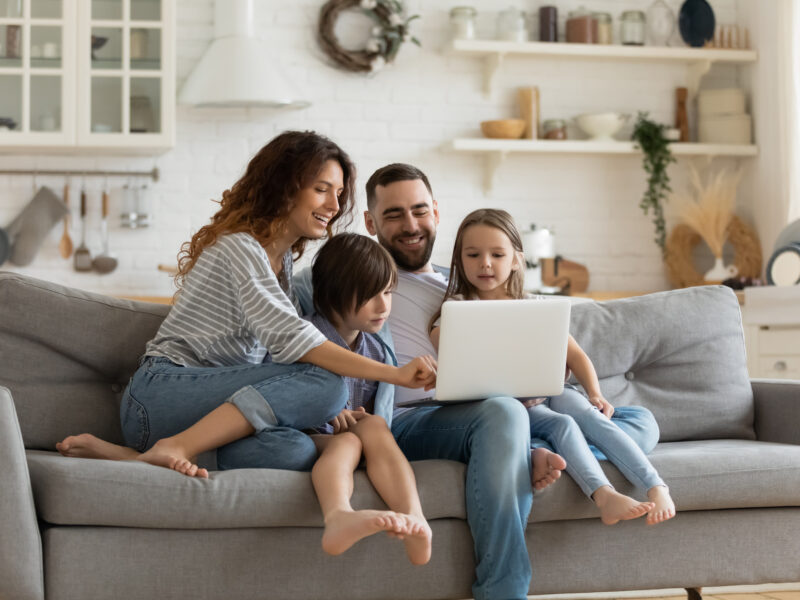 A happy family of four uses a laptop to research open cell vs closed cell insulation while sitting on a gray sofa; white open shelving and walls in the background