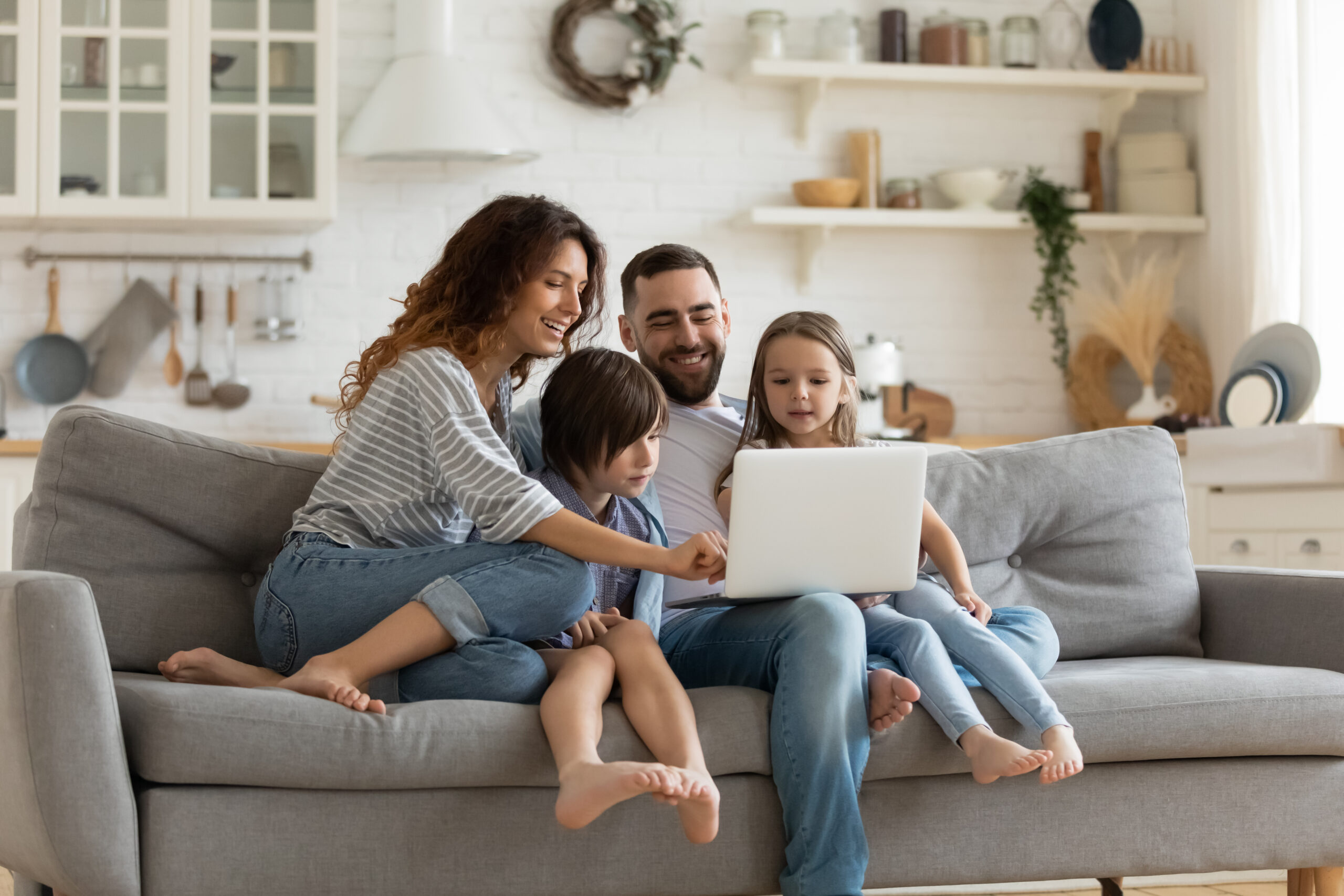 A happy family of four uses a laptop to research open cell vs closed cell insulation while sitting on a gray sofa; white open shelving and walls in the background