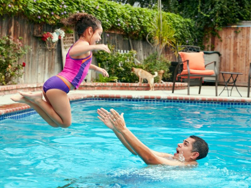 A father catches his daughter who is jumping into their in-ground swimming pool; outdoor seating and a wood fence in the background