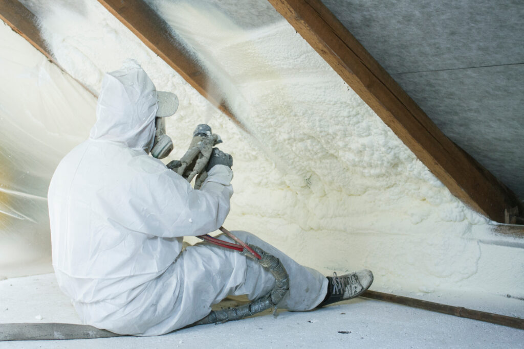 A licensed technician wears white protective gear while applying spray foam insulation to an open attic cavity