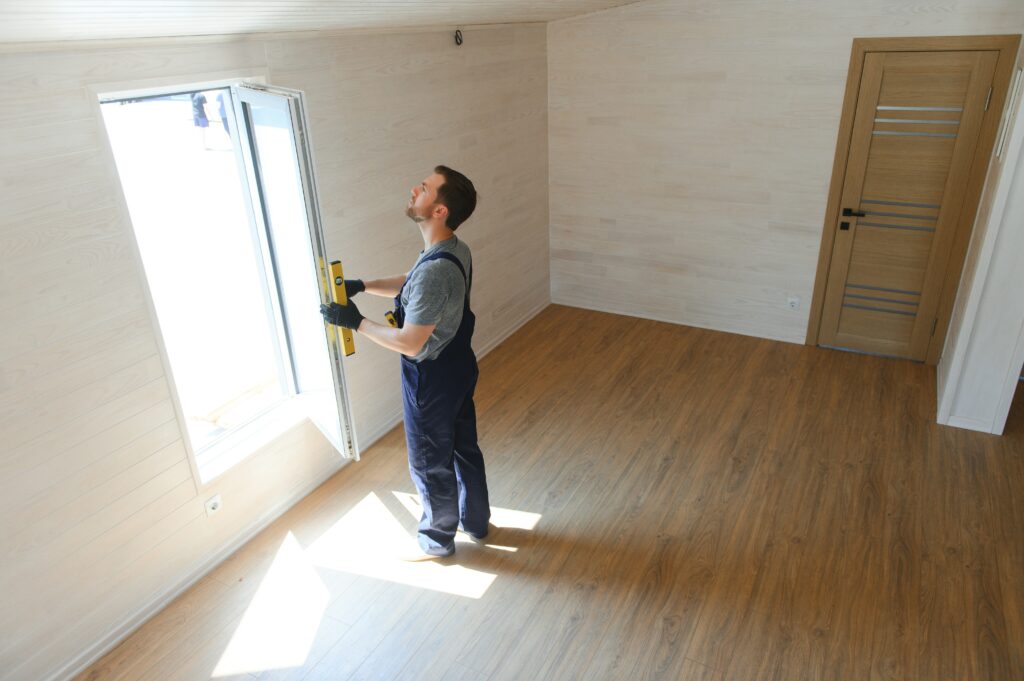 A bearded construction worker in a blue tee shirt and denim overalls inspects a window in a modular home to ensure its proper installation for optimal energy efficiency 