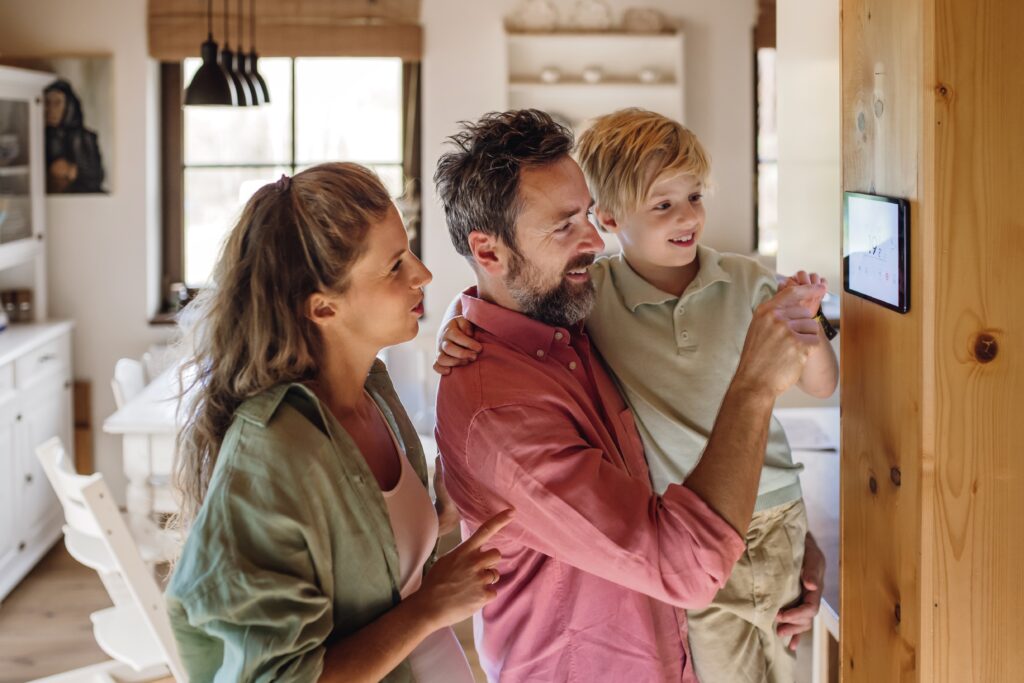 A smiling family looks at the thermostat on a wood wall as they enjoy the energy efficiency of their newly installed spray foam insulation; bright, sunny kitchen in the background  